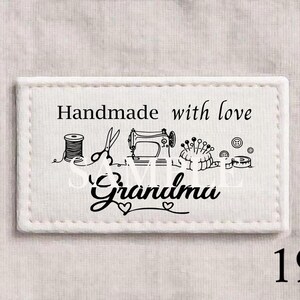 以下が含まれることがあります： 「Handmade with love」の文字と筆記体で書かれた「Grandma」の文字が入った白い長方形の布製ラベル。糸巻き、ハサミ、ミシン、ピン、ボタンなど、縫製道具のイラストが描かれています。