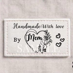 以下が含まれることがあります： 「Handmade With love By Mom SAMPLE」の黒い筆記体とハート型の花の模様が描かれた白い長方形のラベル。デザインの右側には3つの小さなハートの形があります。