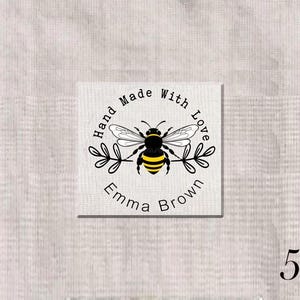 May include: A square white fabric label featuring a black and yellow bee illustration centered between two black leafy branches. The text Hand Made With Love is printed in a black arc above the bee, and the name Emma Brown is printed below.