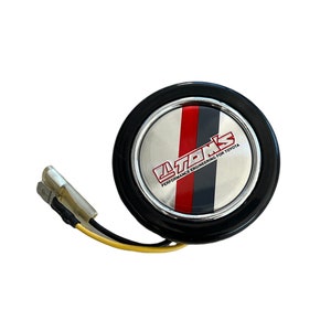 May include: A black steering wheel button with a silver trim and a red and black stripe design. The button has the text "TOM'S PERFORMANCE ENGINEERING FOR TOYOTA" printed on it.