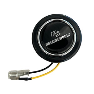 May include: A black and silver MazdaSpeed horn button with the text "MAZDASPEED" in silver lettering. The button has a glossy finish and a silver trim. It includes a connector with black and yellow wires.