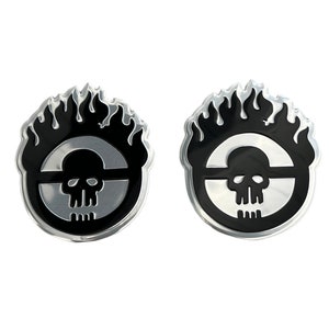 May include: Two black and silver metal car decals featuring a skull with flames behind it. The skull is inside a circle with a line through it.