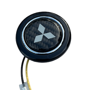 May include: Black carbon fiber steering wheel button with a silver Mitsubishi logo and a black and yellow wire.
