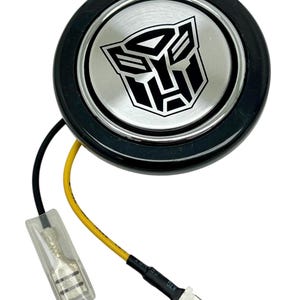 May include: A black round button with a silver center featuring the Transformers logo. The button has two wires attached to it, one yellow and one black.