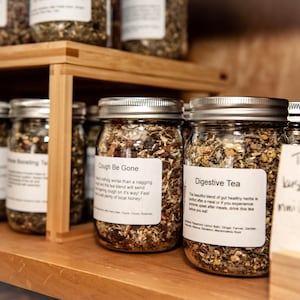 May include: Glass jars filled with dried herbs and teas, labeled with names like "Cough Be Gone" and "Digestive Tea." The jars are arranged on wooden shelves, with a handwritten sign indicating sizes. The tea blends appear to be a mix of green, brown, and tan.
