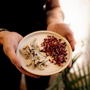 May include: A small, round, white ceramic plate held in hands, divided into two sections. One side holds dried sage, the other holds dried red rose petals. The person holding the plate is wearing a black long-sleeved shirt and a beaded bracelet.
