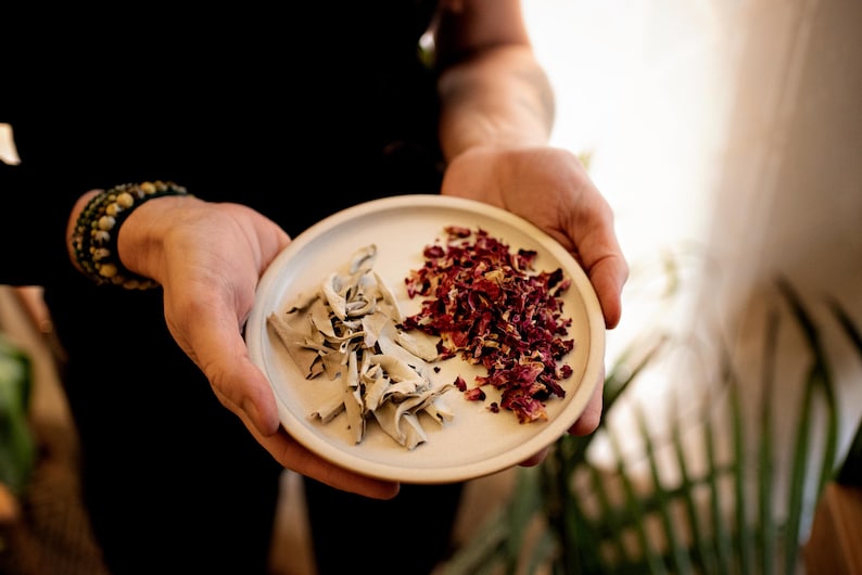May include: A small, round, white ceramic plate held in hands, divided into two sections. One side holds dried sage, the other holds dried red rose petals. The person holding the plate is wearing a black long-sleeved shirt and a beaded bracelet.