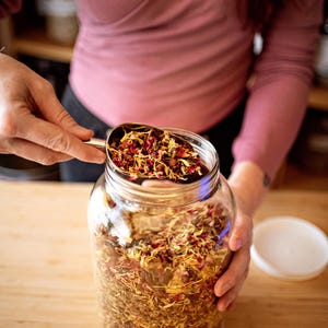 May include: A clear glass jar filled with dried herbs and flowers. A metal scoop holds more of the colorful mixture, with red and yellow petals visible. The jar sits on a wooden surface, with a white lid nearby.