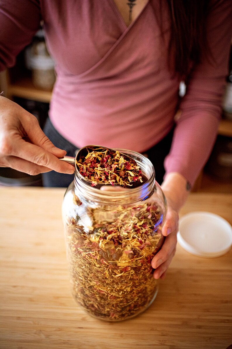 May include: A clear glass jar filled with dried herbs and flowers. A metal scoop holds more of the colorful mixture, with red and yellow petals visible. The jar sits on a wooden surface, with a white lid nearby.