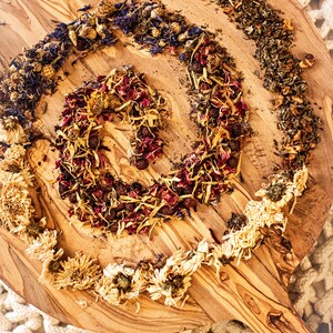 May include: A wooden cutting board with a spiral arrangement of dried flowers, herbs, and berries. The arrangement includes dried yellow flowers, blue petals, and red berries. The board has a natural wood grain pattern.