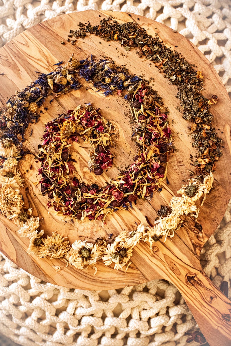 May include: A wooden cutting board with a spiral arrangement of dried flowers, herbs, and berries. The arrangement includes dried yellow flowers, blue petals, and red berries. The board has a natural wood grain pattern.
