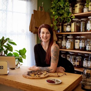 May include: A woman stands behind a wooden counter with a Square payment system and a cutting board with food. Shelves filled with jars of herbs and spices are in the background. Plants and a clear glass kettle are also visible.
