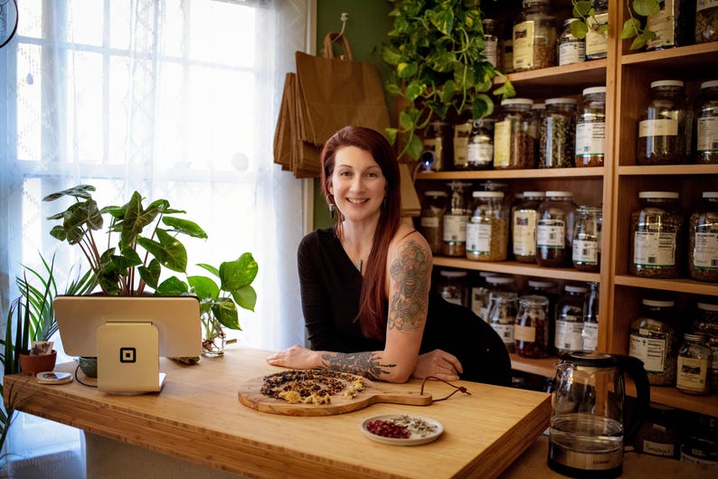 May include: A woman stands behind a wooden counter with a Square payment system and a cutting board with food. Shelves filled with jars of herbs and spices are in the background. Plants and a clear glass kettle are also visible.