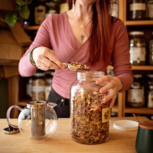 May include: A woman using a scoop to fill a glass jar with dried herbs and flowers. A clear glass teapot with a metal infuser sits on the wooden counter. Shelves of jars are in the background.