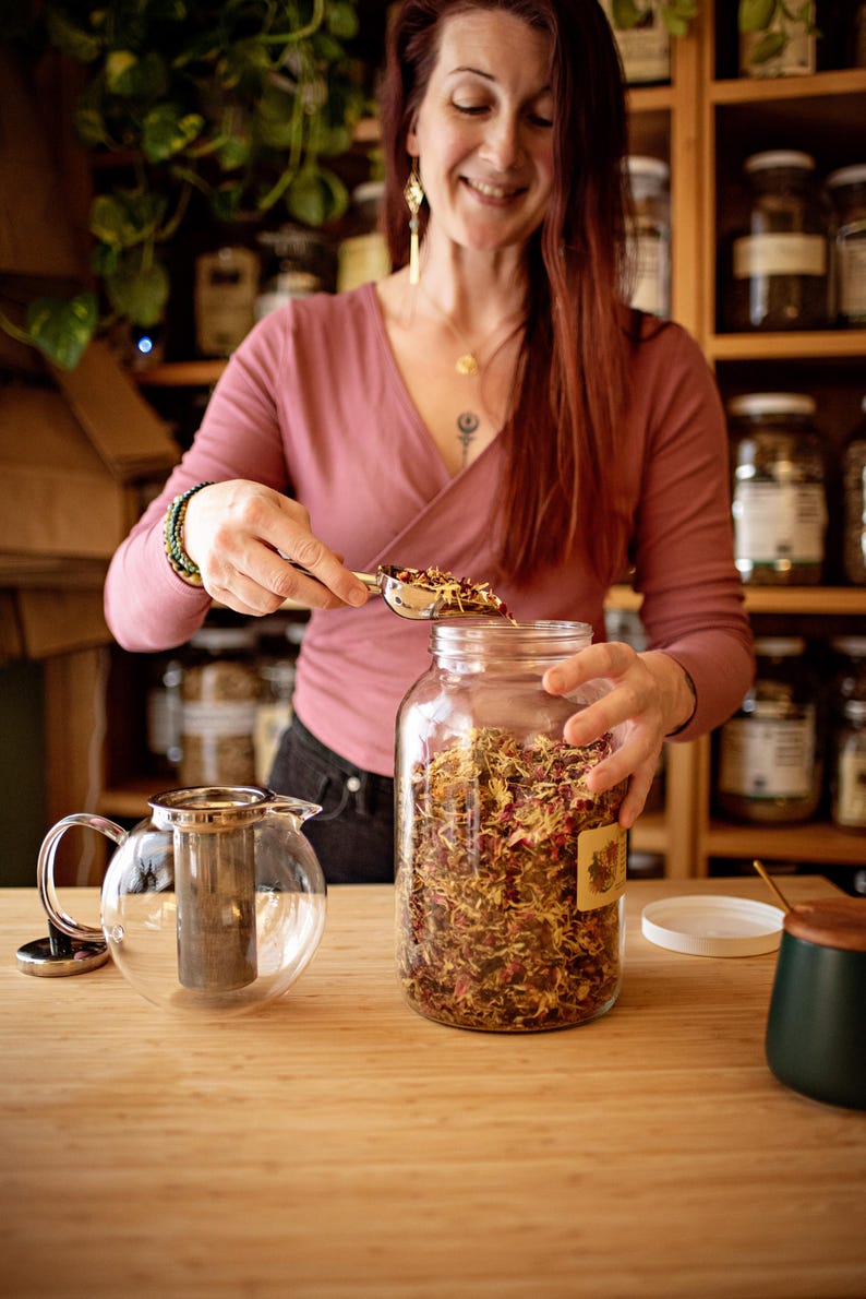 May include: A woman using a scoop to fill a glass jar with dried herbs and flowers. A clear glass teapot with a metal infuser sits on the wooden counter. Shelves of jars are in the background.