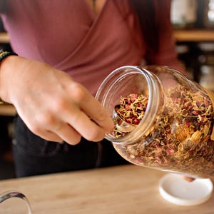 May include: A clear glass jar filled with dried herbs and flowers is being held. A silver spoon is scooping out the contents. The jar has a brown label. A person wearing a pink shirt and bracelets is holding the jar.