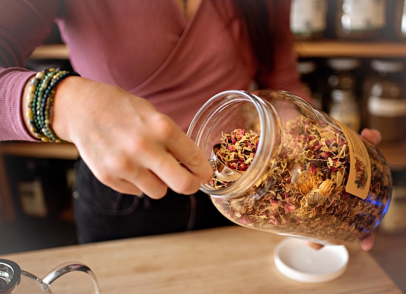 May include: A clear glass jar filled with dried herbs and flowers is being held. A silver spoon is scooping out the contents. The jar has a brown label. A person wearing a pink shirt and bracelets is holding the jar.