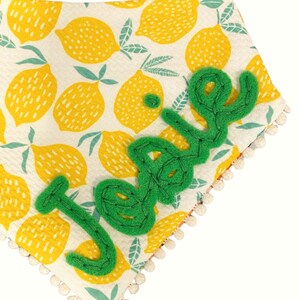 May include: A white bib with a lemon print and a green felt name "Josie" sewn on. The bib has a white pom-pom trim.
