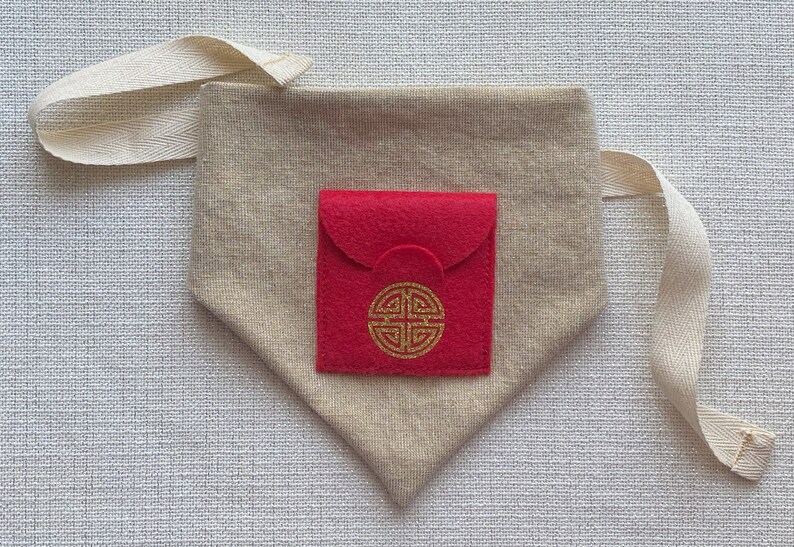 Pu&ograve; includere: Una piccola borsa in feltro rosso con un simbolo dorato sul davanti, all'interno di una borsa in tessuto beige pi&ugrave; grande con una tracolla bianca.