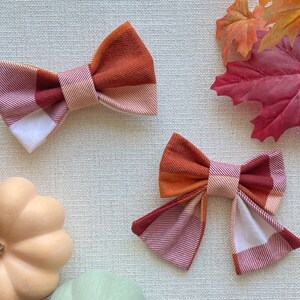 May include: Two plaid fabric hair bows in shades of orange, red, and white. The bows are tied with a white ribbon and are perfect for adding a touch of autumn style to any outfit.