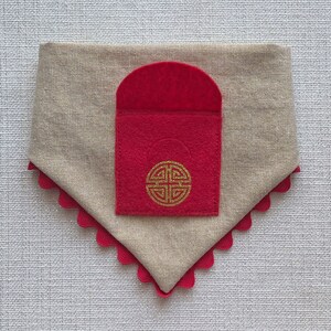 Pu&ograve; includere: Un bandana per cani in lino beige con una tasca in feltro rosso. La tasca ha un disegno di nodo cinese dorato.