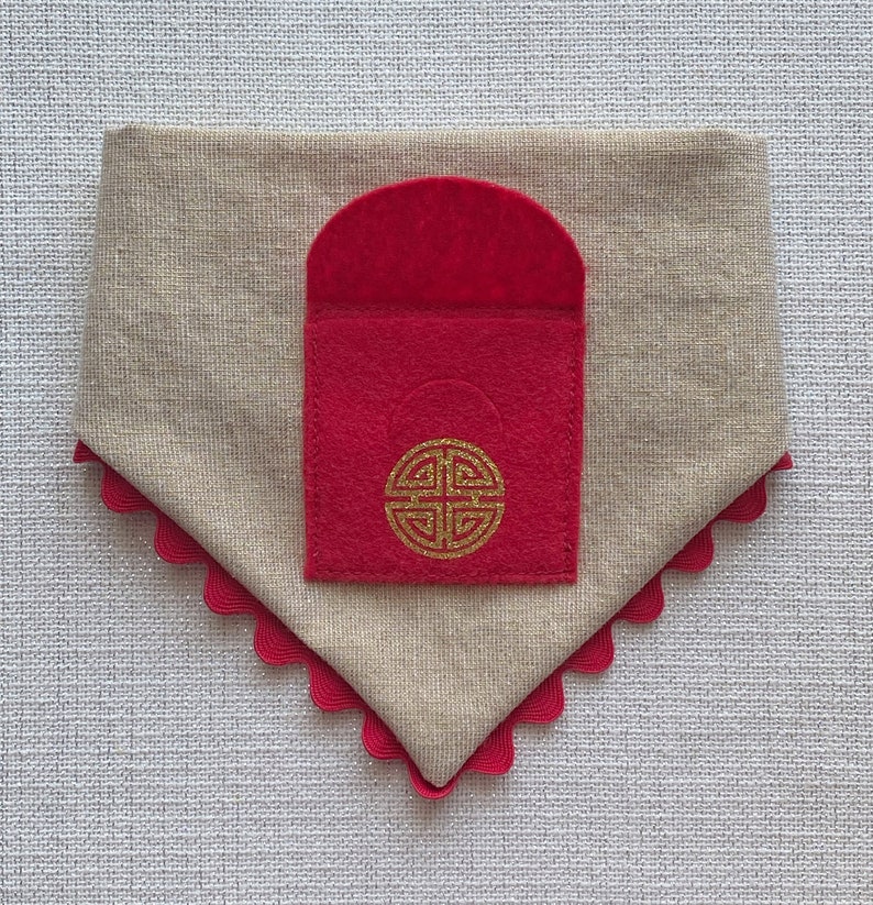 Pu&ograve; includere: Un bandana per cani in lino beige con una tasca in feltro rosso. La tasca ha un disegno di nodo cinese dorato.