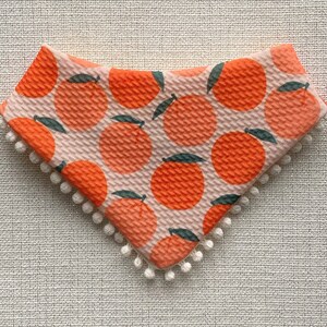 May include: A pink and orange fabric bib with a white pom-pom trim. The bib features a repeating pattern of orange slices with green leaves.
