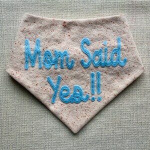Puede incluir: Un babero de fieltro marr&oacute;n claro con texto bordado en azul que dice "Mom Said Yes!!"