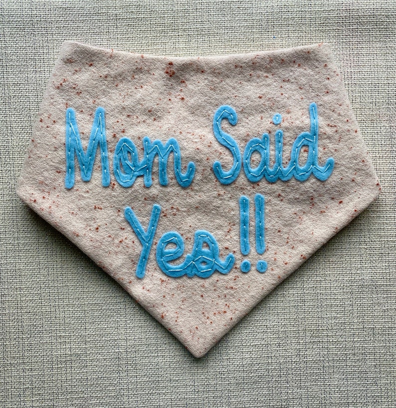 Puede incluir: Un babero de fieltro marr&oacute;n claro con texto bordado en azul que dice "Mom Said Yes!!"