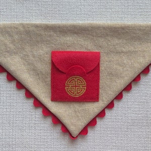 Pu&ograve; includere: Un bandana per cani in tessuto beige con una tasca in feltro rosso. La tasca ha un disegno di nodo cinese dorato.