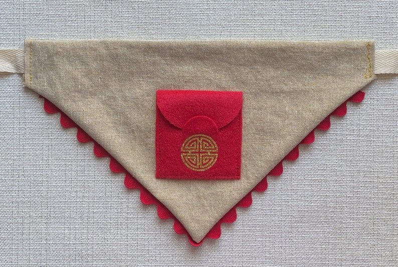 Pu&ograve; includere: Un bandana per cani in tessuto beige con una tasca in feltro rosso. La tasca ha un disegno di nodo cinese dorato.