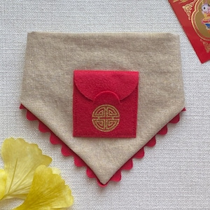 Pu&ograve; includere: Un bandana per cani in tessuto beige con una tasca in feltro rosso e un design a nodo cinese dorato. Il bandana ha un bordo smerlato.
