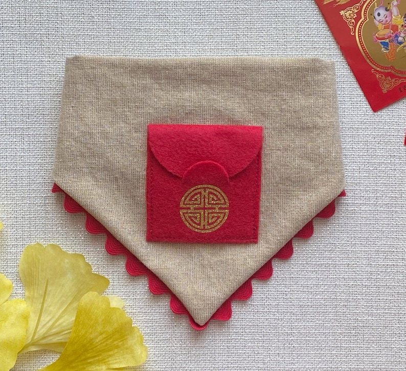 Pu&ograve; includere: Un bandana per cani in tessuto beige con una tasca in feltro rosso e un design a nodo cinese dorato. Il bandana ha un bordo smerlato.