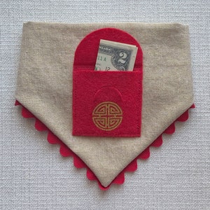 Pu&ograve; includere: Una busta in feltro rosso con un disegno di nodo cinese dorato e un biglietto da 2 euro all'interno. La busta &egrave; attaccata a un tessuto beige con un bordo a festoni rosso.
