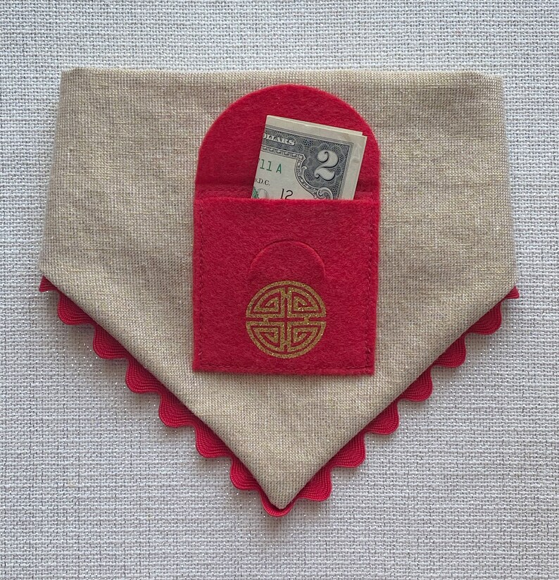Pu&ograve; includere: Una busta in feltro rosso con un disegno di nodo cinese dorato e un biglietto da 2 euro all'interno. La busta &egrave; attaccata a un tessuto beige con un bordo a festoni rosso.