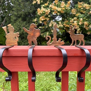 May include: A set of four metal stocking holders featuring brass-colored silhouettes of a snowman, Santa Claus, a candle, and a reindeer. Each figure is attached to a black, curved metal hook designed to hang over the edge of a flat surface.