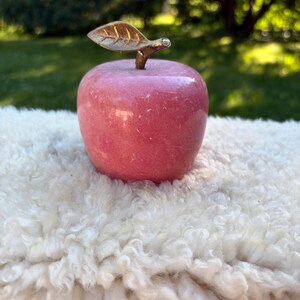 May include: A polished pink stone apple figurine featuring a smooth, rounded body and a textured metallic gold-toned stem with a single leaf attached to the top. The object has a glossy finish and a solid, uniform appearance throughout its surface.