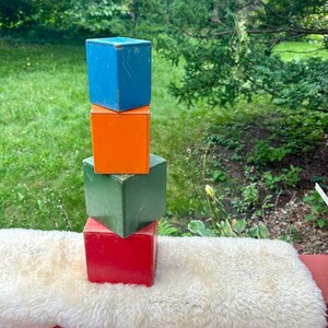 May include: A stack of four wooden blocks in different colours. The bottom block is red, the next is green, the next is orange, and the top block is blue.