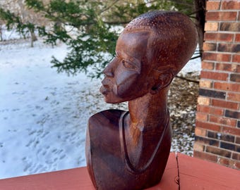 May include: A carved wooden bust of a person with a smooth, rounded head and detailed facial features. The sculpture is a rich, dark brown color and is positioned on a red wooden railing. The background includes trees, snow, and a brick wall.
