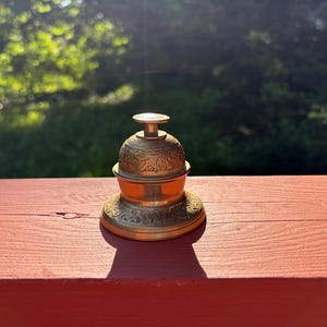 May include: A brass desk bell featuring an intricate embossed floral pattern on the dome and base. The bell has a cylindrical push button at the top and a wide, circular foundation, showcasing a metallic gold finish with visible light reflections.