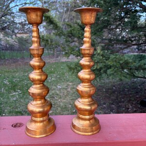 May include: A pair of tall, metallic gold-toned candle holders featuring a stacked, multi-tiered design with a wide circular base, a slender central column composed of several rounded, bulbous sections, and a flared top rim for holding candles.