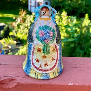 May include: A ceramic bell figurine depicting a woman in traditional attire, holding a bouquet of flowers. The bell features a blue head covering with red dots, a yellow apron with floral accents, and a blue and yellow dress. The background is blurred.