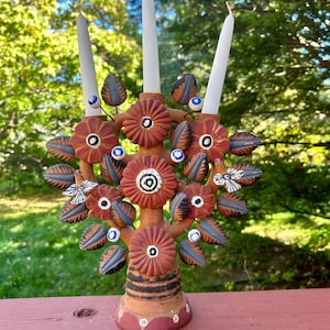 May include: A handcrafted, terracotta candle holder shaped like a tree, adorned with flowers, leaves, and small blue accents. It holds three white candles. The base has a striped design and is set on a red surface.