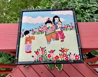 May include: Needlepoint artwork featuring three figures in traditional attire amidst a field of flowers. The figures are rendered in vibrant colors, set against a sky blue backdrop with fluffy white clouds. The artwork is framed in a black bamboo-style frame.