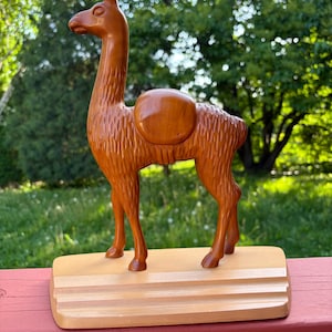 May include: A carved wooden llama figurine with a smooth, polished reddish-brown finish and textured fur detailing. The llama features a rounded saddle on its back and stands on a light-colored, rectangular wooden base with tiered edges.