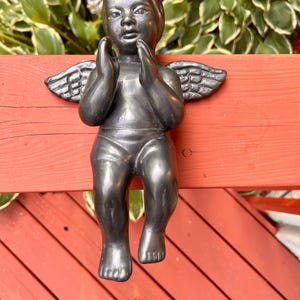 May include: A dark gray cherub statue with small wings, hands clasped near its face, and seated legs. The statue is mounted on a red wooden surface, with green foliage in the background. The cherub is a decorative garden ornament.