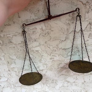 May include: A vintage hanging balance scale featuring a dark metal horizontal beam and a central suspension hook. Two circular brass pans are suspended from the ends of the beam by thin metal chains, creating a symmetrical weighing instrument.