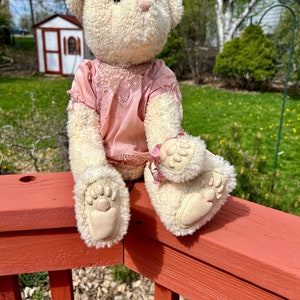 May include: A white plush teddy bear wearing a pink lace dress is sitting on a red wooden railing. The bear is looking up and has a sweet expression on its face.