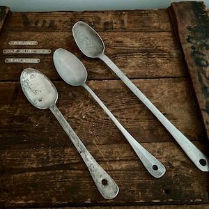 May include: Three silver metal spoons of varying sizes on a wooden surface. The spoons have a rounded bowl and a long handle.