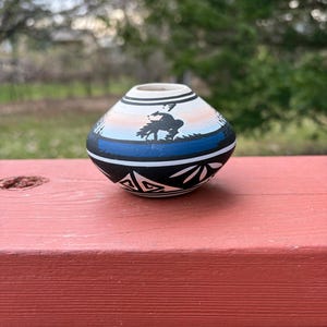May include: A small, round ceramic pot featuring a hand-painted black silhouette of a bucking horse and rider. The vessel has a white rim, a light orange and blue gradient band, and a black base decorated with white geometric leaf patterns.